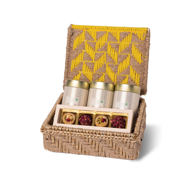 Woven gift basket with yellow patterned lid containing tea canisters and small items on a white background